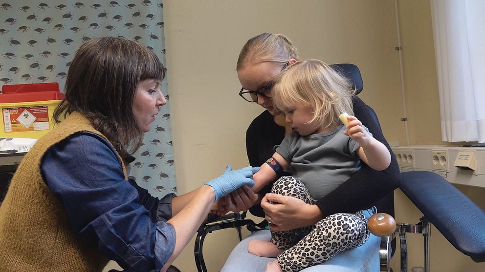 En sjuksköterska letar blodkärl på tvååring som sitter i sin mammas knä.