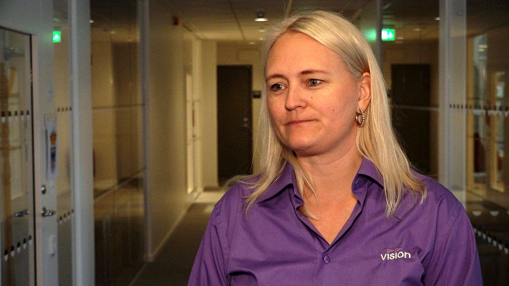 Kvinna i lila skjorta står i en kontorskorridor med glasväggar och stängda dörrar.