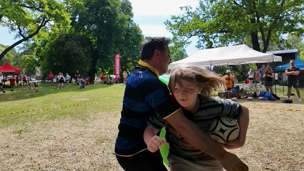 En man i mörka kläder hindrar en pojke att ta sig fram. Pojken håller en rugbyboll