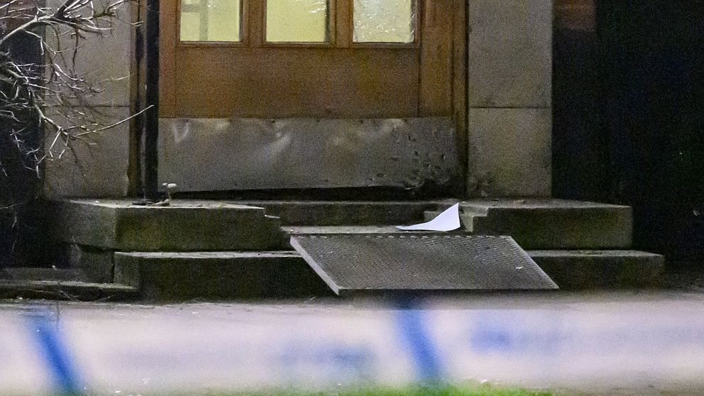 En trappa med en trasig mattläggning framför en dörr, omgiven av polisavspärrning. En vit pappersbit ligger på trappan, och det syns också några avblommade kvistar i närheten.
