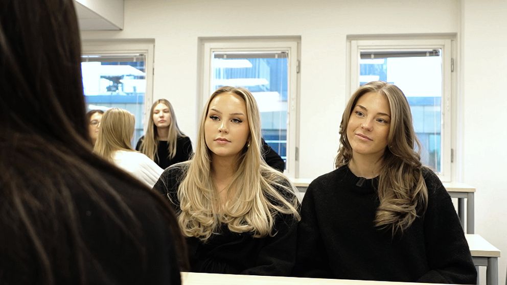 Tre personer i svarta tröjor sitter vid ett bord i ett ljust rum. Den främre personen syns delvis i bild och riktar sig mot de två andra, som har långt hår och ser mot kamera. I bakgrunden syns en del av en grupp kvinnor som också sitter. Rummets design är modern med stora fönster som släpper in mycket ljus.