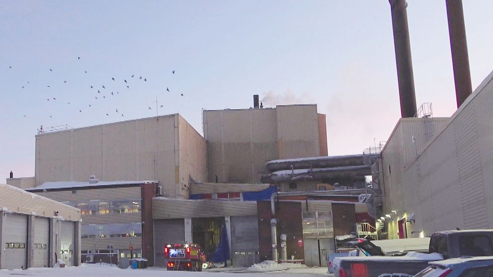 Värmeverk med rök som stiger från skorstenar, omgiven av snö. En brandbil är parkerad framför byggnaden, och fåglar flyger i luften ovanför.
