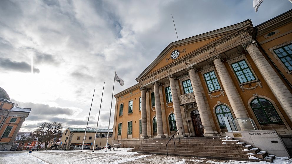 Stadshusets fasad med pelare och klocka, omgiven av snötäckt mark och ett dramatiskt molnigt sky. Flaggar är hissade på stång framför byggnaden.