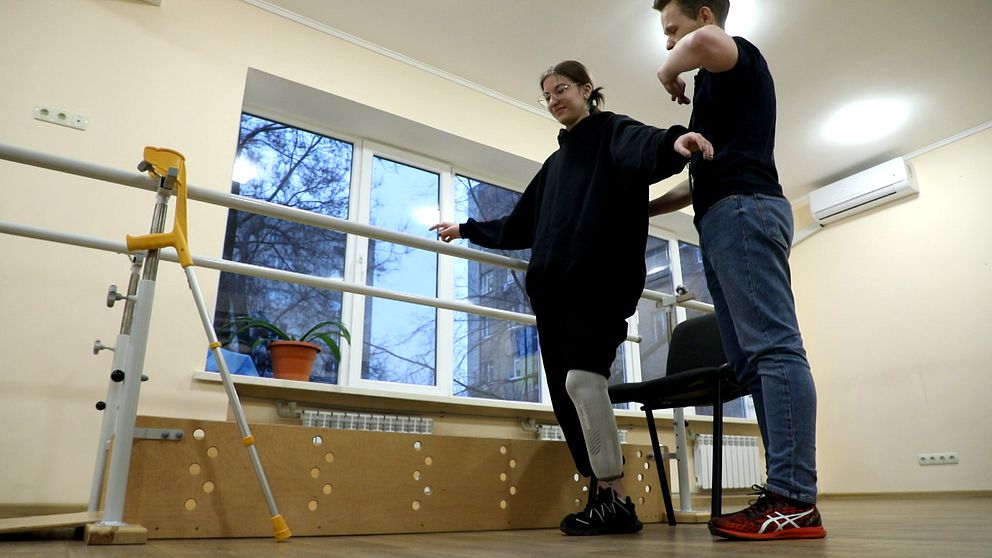 En person med protes står vid en balansstav med stöd av en annan person, i en rehabiliteringsmiljö.