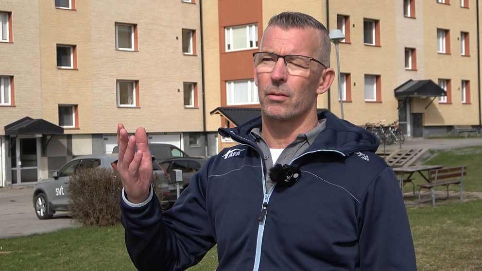 Man i blå jacka står utomhus framför ett flerfamiljshus, där AI styr inomhustemperaturen, och gör en gest med handen.