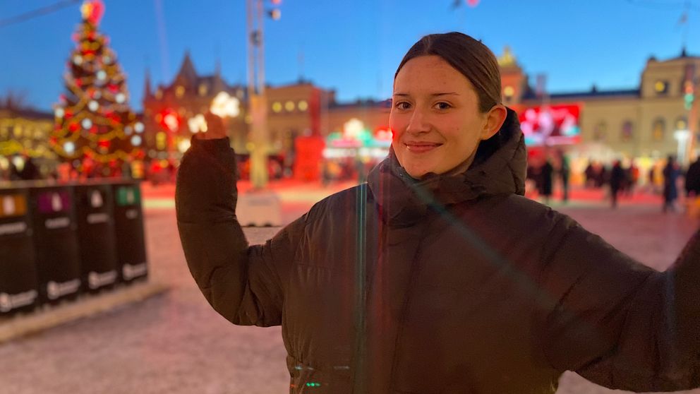 En person i en varm, svart jacka står utomhus med armarna uppsträckta, omgiven av en festlig atmosfär med juldekorationer. I bakgrunden syns en julgran och ljus som pryder området, med byggnader som har festlig belysning. Scenen är upplyst av kvällsljus i en vintermiljö.