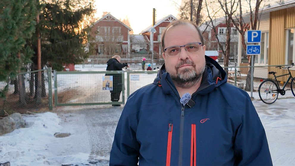 En person i mörk jacka står vid en grind som leder in till en snötäckt väg. I bakgrunden syns en parkering med en cykel och en person som går mot en annan som står vid en bänk. Området är omgiven av träd och hus, vilket skapar en vinterlig atmosfär. Solen går ner i horisonten, vilket ger en varm ton till bilden.