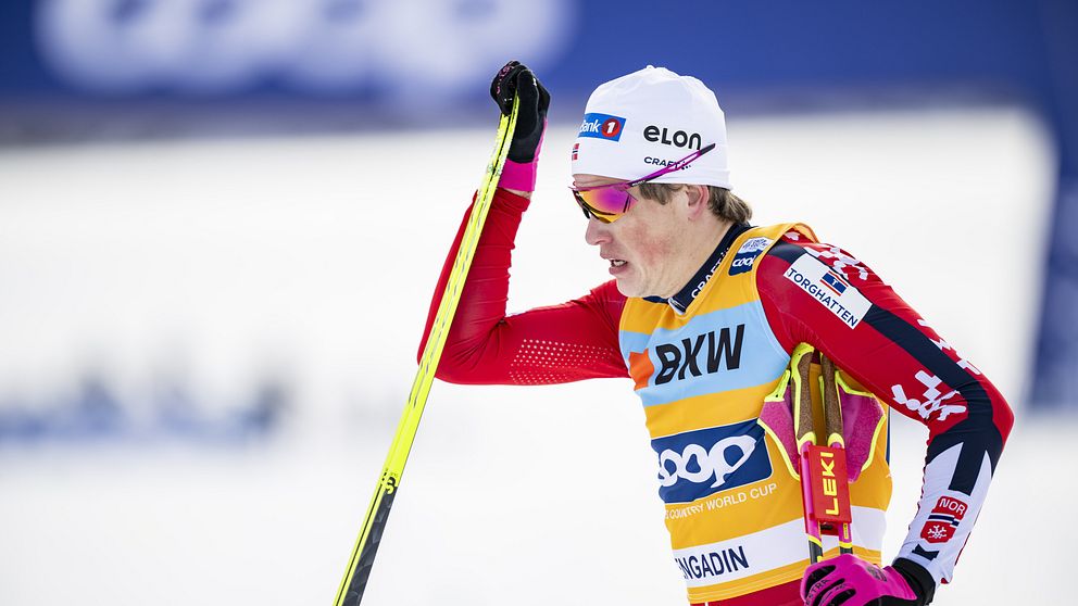 En längdskidåkare klädd i röd tävlingsdräkt med sponsorsloggor lutar sig mot en skidstav. Skidåkaren bär gula och röda handskar, en vit mössa med sponsormärken och en gul tävlingsväst märkt ”COOP FIS World Cup” samt ”ENGADIN”. Bakgrunden är snötäckt med en suddig banderoll med texten ”COOP”.