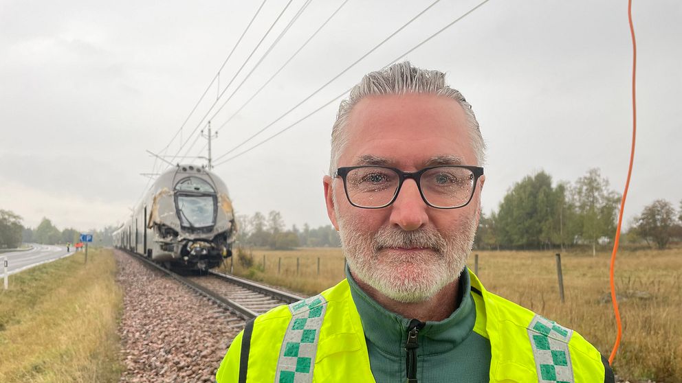 En man med reflexväst står framför ett tåg som är förstört efter en krock.
