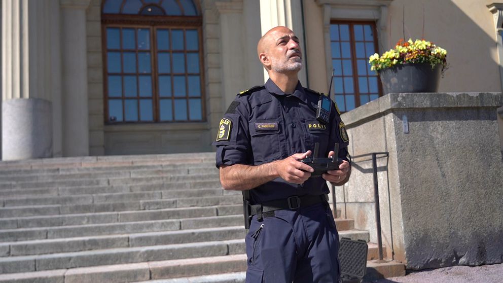 En uniformerad polis står framför en stentrappa med en handkontroll. Har blicken mot skyn.