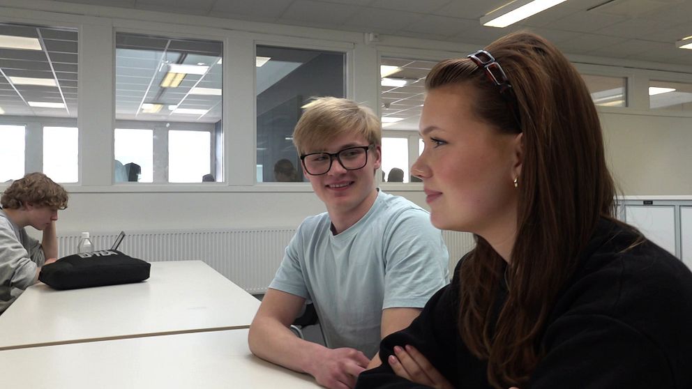 Två personer sitter vid ett bord i ett ljust klassrum eller biblioteksmiljö.
