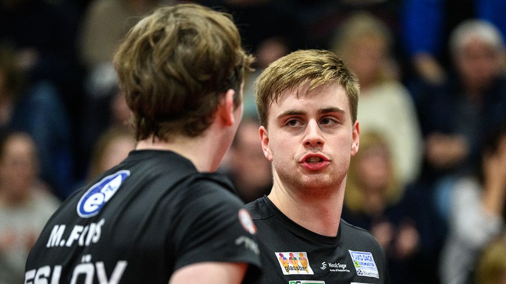 Två bordtennisspelare i svarta tröjor samtalar under en match inför publik.