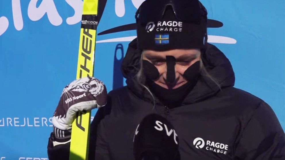 En person i skidsportutrustning står framför en blå bakgrund med logotyper. Personen håller en skida och har på sig en svart jacka med sponsorlogotypen Ragde Charge. Det ser ut att vara en intervju efter en tävling.