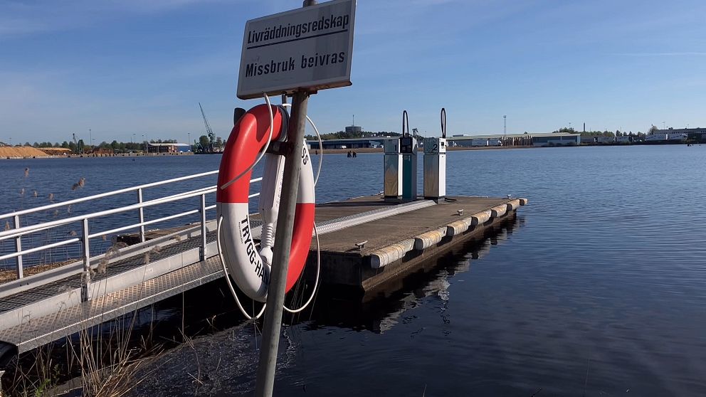 I förgrunden syns en röd och vit räddningsboj på en stolpe och i bakgrunden en sjömack med två pumpar på en brygga i Vänern.