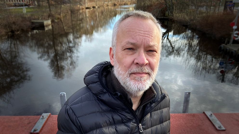 En person i svart jacka står på en bro med en spegelblanka sjö i bakgrunden, omgiven av träd och en lugn landskapsvy.
