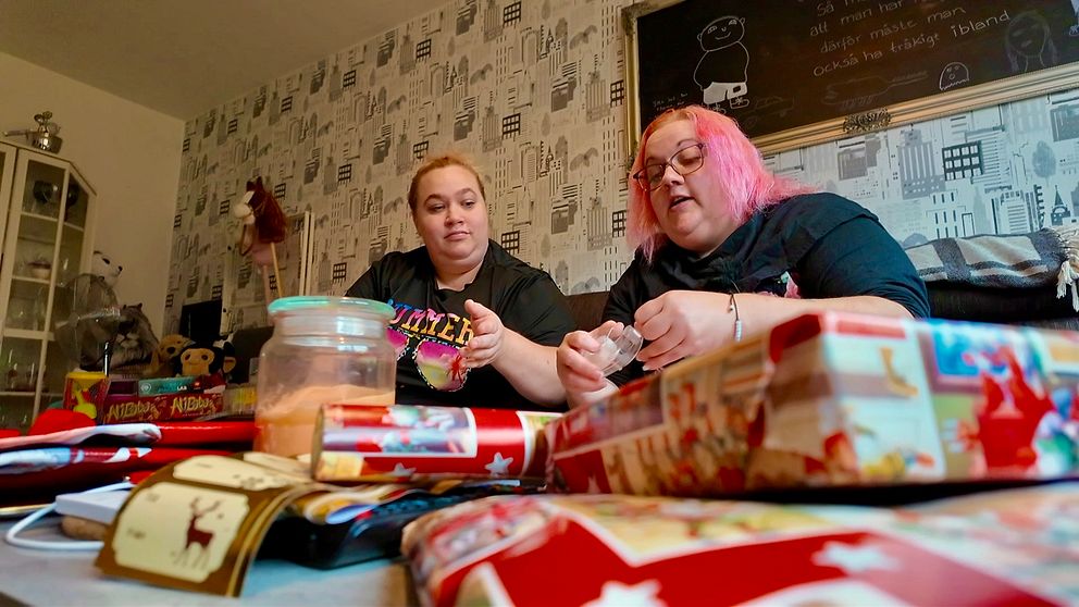 Två personer sitter vid ett bord och packar in julklappar. Framför dem ligger olika inslagna paket och dekorationer, inklusive ett glas med något i och rullar av presentpapper. I bakgrunden syns en vägg med tapetdesign och en tavla med text. Atmosfären är hemtrevlig och festlig, vilket antyder att de förbereder sig för en högtid.