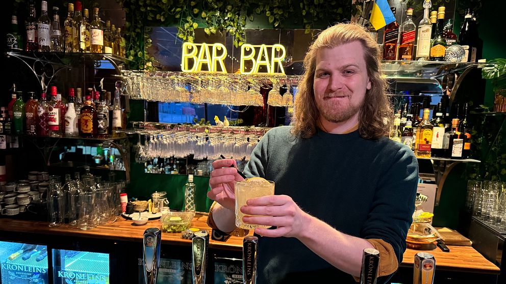 Bartender serverar en drink i en livlig bar med flaskor och glas i bakgrunden.