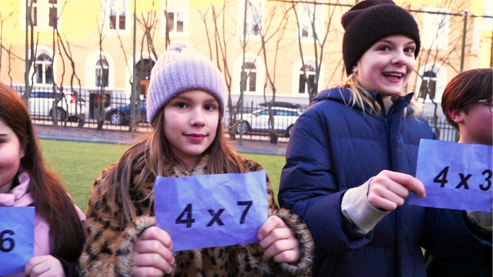 Barn håller upp färgglada kort med matematiska multiplikationsuppgifter utomhus. Tre av dem visar talen ”4 x 7”, ”6” och ”4 x 3”. Bakar och klädstilen tyder på en kall väderlek.