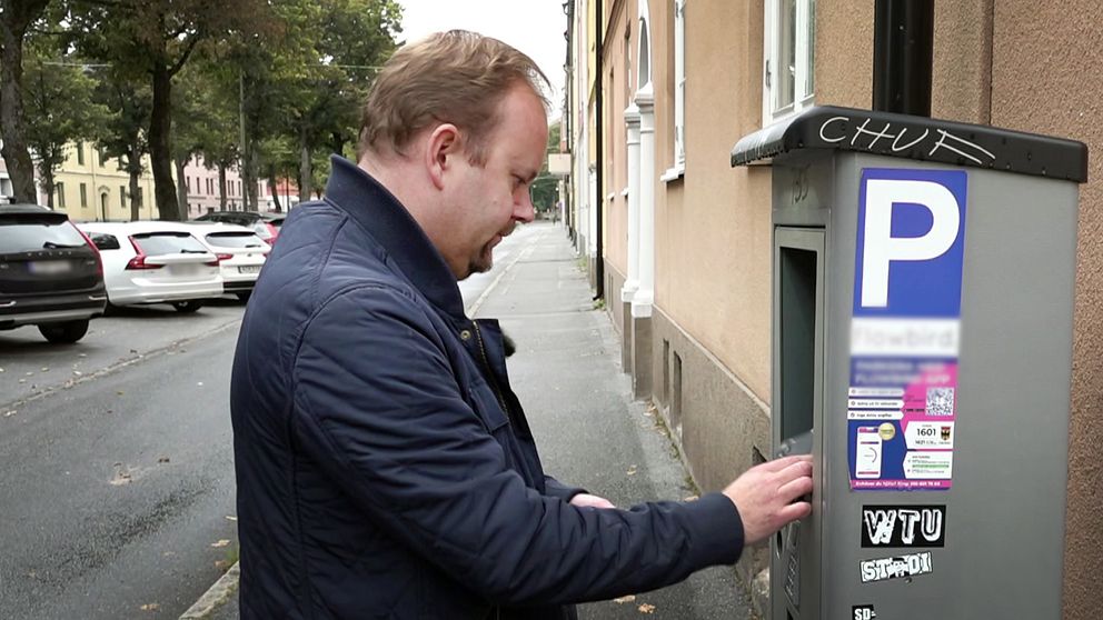 En person står vid en parkeringsautomat och använder den, med ett parkeringstecken synligt på automaten. I bakgrunden syns en rad med parkerade bilar och en gata kantad av träd.