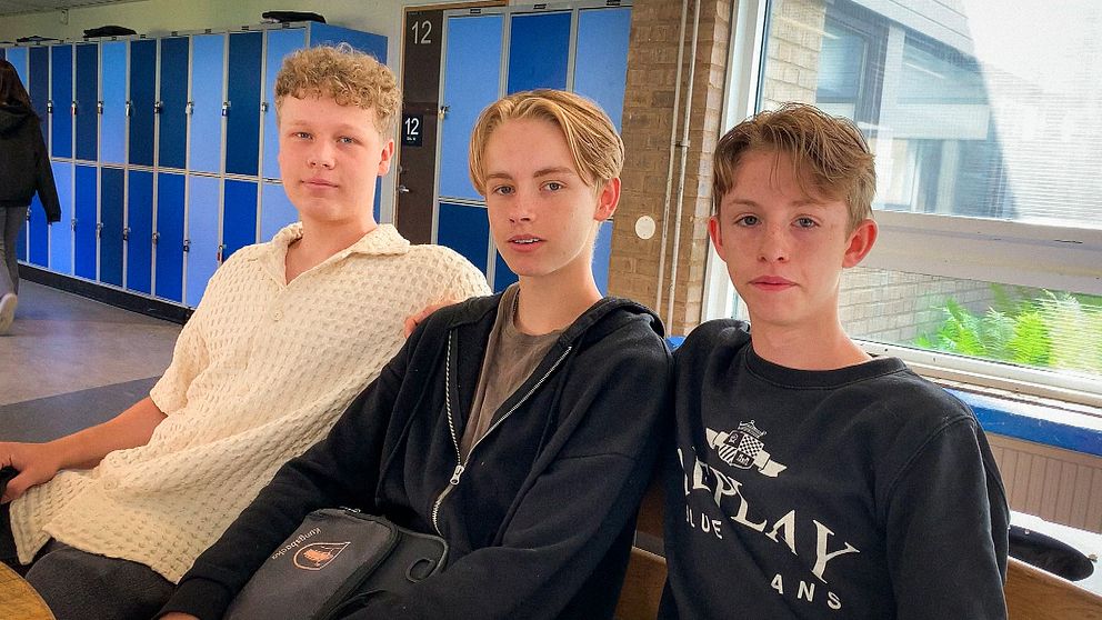 Tre killar sitter på en bänk i en skolkorridor.