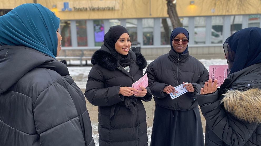 Fyra kvinnor i vinterkläder står utomhus och samtalar, några av dem håller papper i händerna. I bakgrunden syns en gul byggnad med fönster.