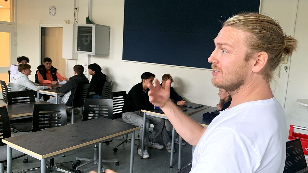 Samhällskunskaps läraren Daniel Skarp står och gestikulerar medan eleverna i klassrummet sitter i grupper runt två olika bord och diskuterar.