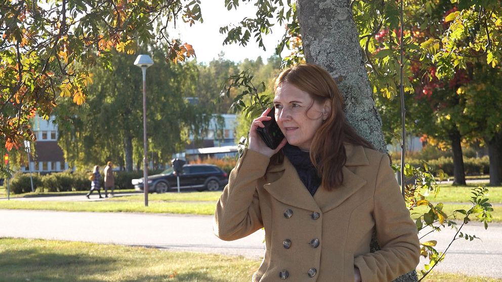 En person står lutad mot ett träd och pratar i telefon. Bakgrunden visar en park med grönska och fler människor som promenerar. Det är en solig dag med klar himmel och höstlöv i olika färger.