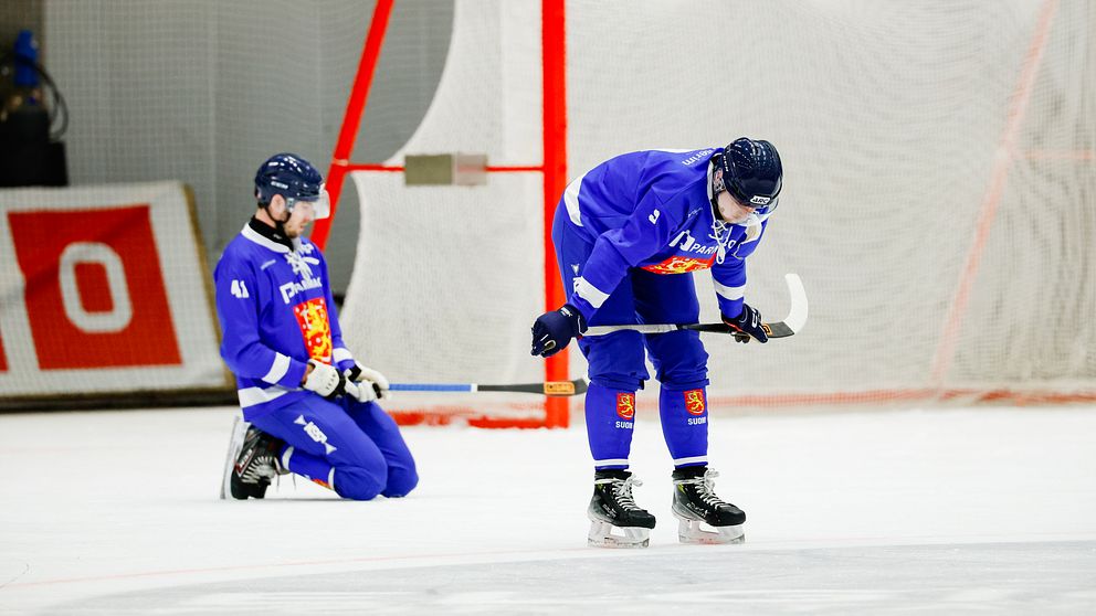 Två bandyspelare i blåa dräkter ser besvikna ut framför ett mål på isen.