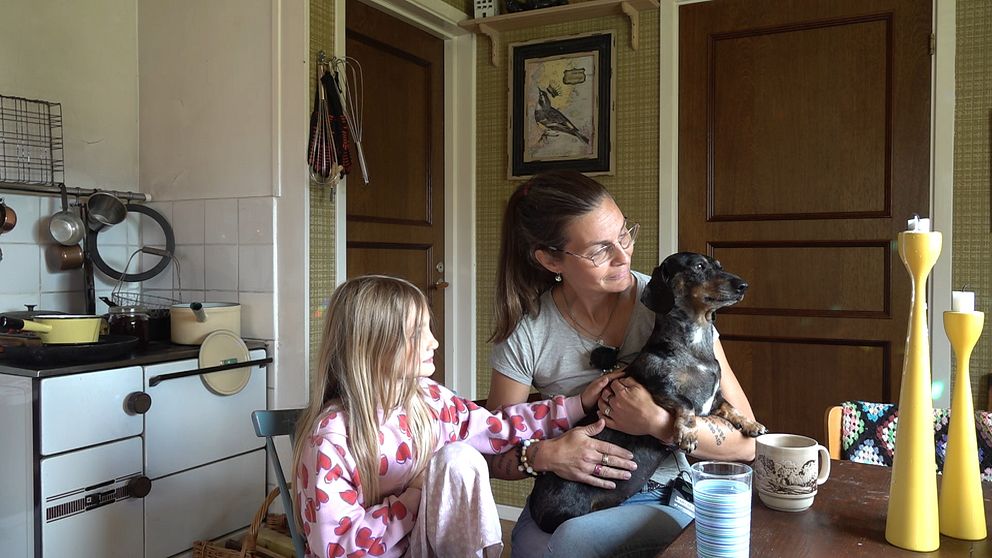 I ett 70-tals kök vid borde sitter en liten tjej och hennes mamma som har en tax-hund i knät