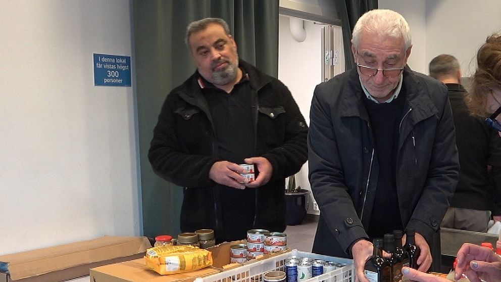 Två män i svarta jackor packar upp matvaror på ett bord. En kvinna med telefon köper en vara.
