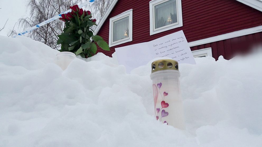 Ett minnesmärke i snön utanför ett rött hus. Märket består av ett blockljus dekorerat med hjärtan, en bukett röda rosor och ett handskrivet brev.