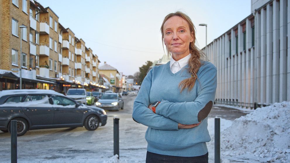 En person står med armarna i kors på en gata i en stad, omgiven av snö. I bakgrunden syns flerbostadshus och flera bilar parkerade längs gatan.