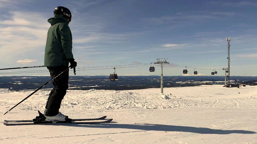 En skidåkare i grön jacka står på en snötäckt backe och blickar mot en fjärran utsikt, medan skidliftar, gondoler, svävar över landskapet i bakgrunden.