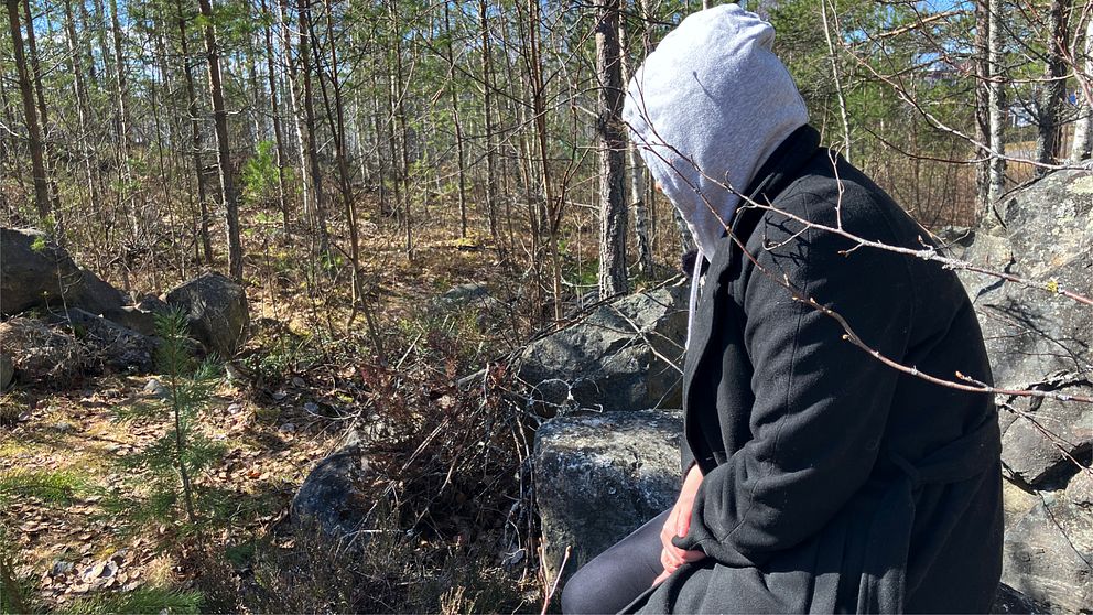 En tjej sitter med uppdragen huva och svart kappa på en sten i skogen