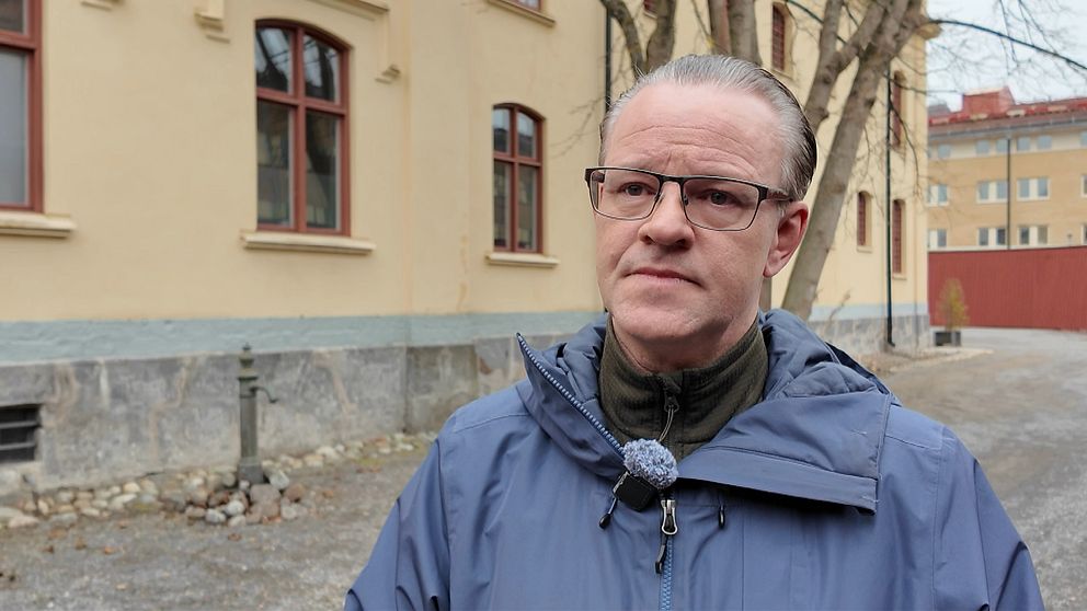 Person utomhus framför gulputsad byggnad med röda fönster och träd i bakgrunden.