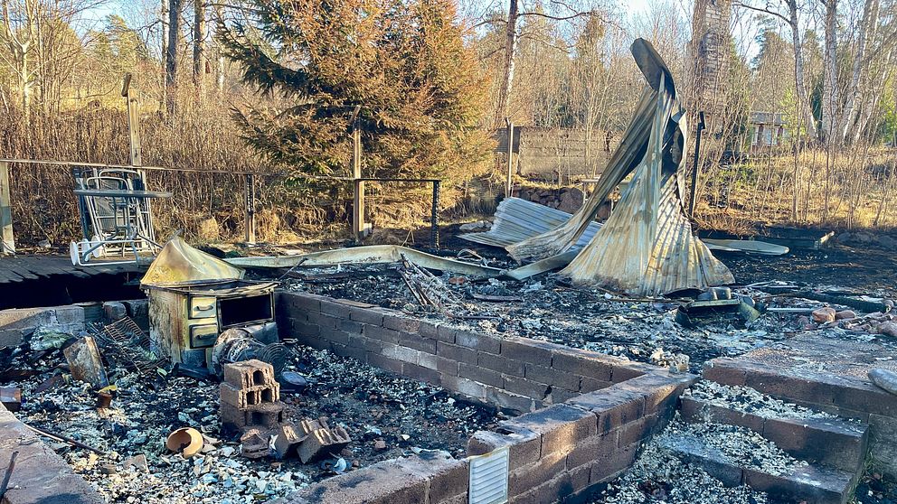 Rester av en utbränd byggnad på en tomt, med förkolnade tegelväggar och förvridna metallstrukturer. Bakgrunden visar träd och en åker.