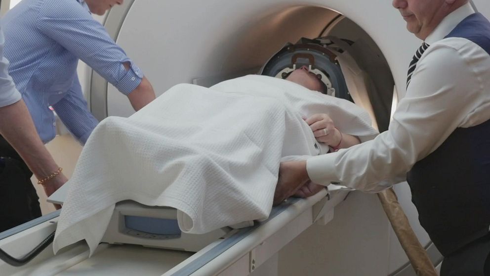 En patient ligger på en bår som förs in i en MRI-maskin, med två vårdpersonal som hjälper till. En person är klädd i en skjorta med lång ärm och en annan i en vit skjorta och mörk väst. Patienten är täckt med en filtar och har en huvudskyddsanordning på sig.