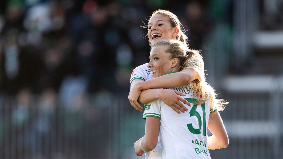 Fotbollsspelare i gröna och vita dräkter firar mål med en kram på spelplanen.
