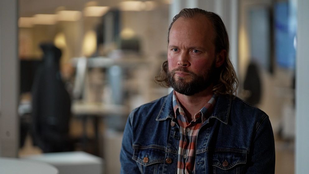 En person sitter framåtlutad i en kontorsmiljö, iförd en jeansjacka och en rutig skjorta. I bakgrunden syns oskarpa kontorsdetaljer och datorer.