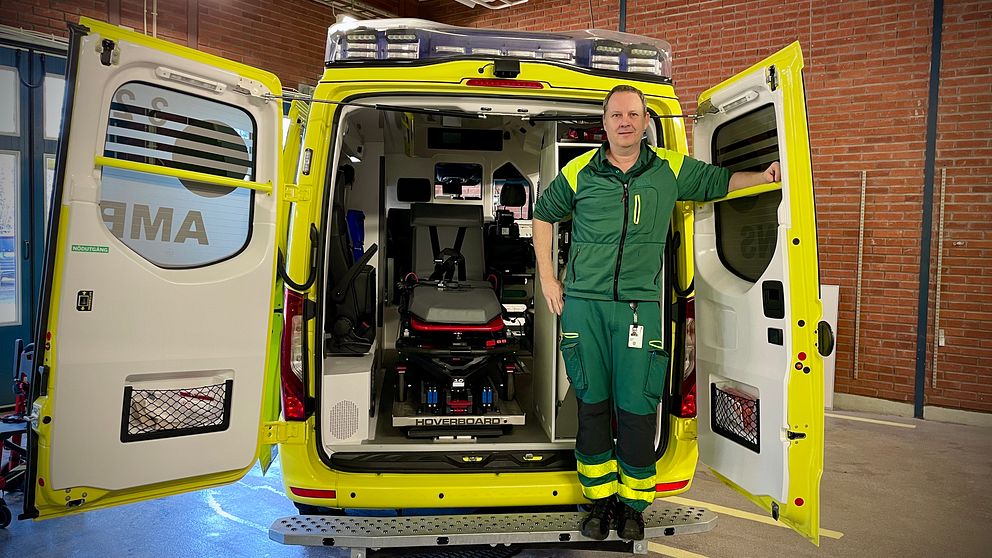 En ambulans med öppen bakdörr visar insidan som har en bår och medicinsk utrustning. En person i gröna arbetskläder står vid ambulansens öppna dörrar.