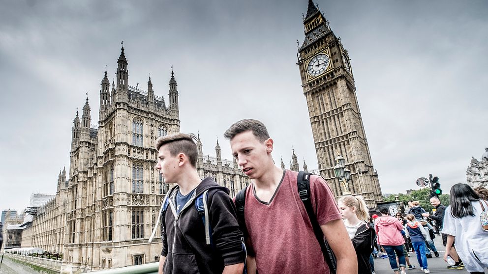 Ung man står framför Big Ben i London. Folk går på bron i bakgrunden.