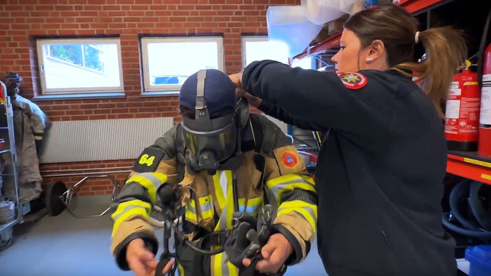 En brandman är på väg att sätta på sig sin skyddsdräkt med hjälp av en medhjälpare. Rummet är inrett med brandutrustning och röda brandsläckare syns i bakgrunden, samt ett par fönster som släpper in dagsljus.