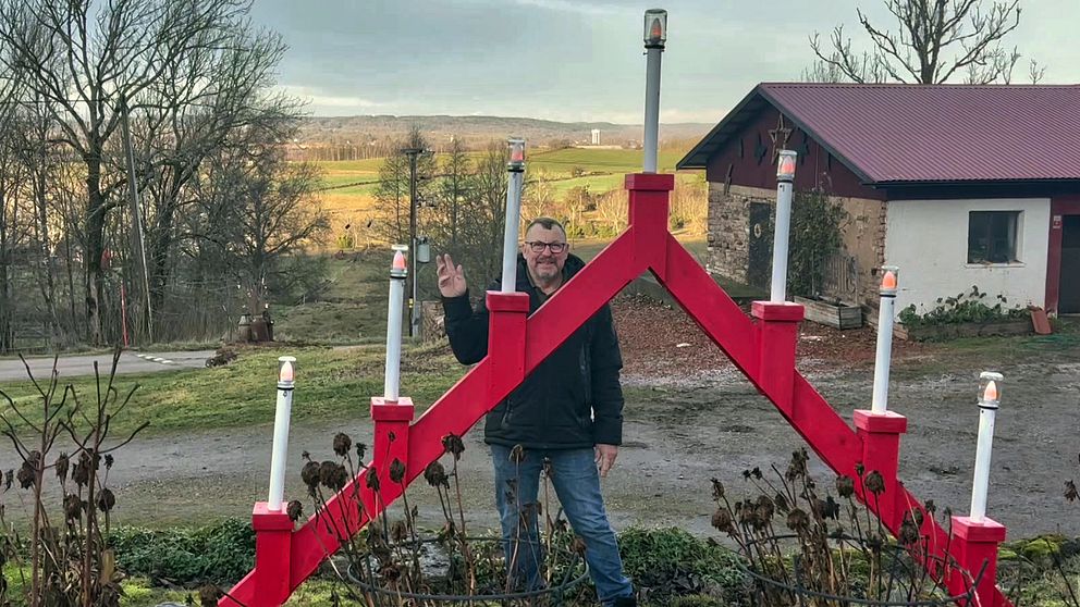 En person står framför en stor, röd dekoration i form av en stor adventsljusstake. I bakgrunden syns en landskapsvy med grönskande ängar och träd, samt en byggnad med röd tak. Vädret ser klart ut med en ljus himmel.