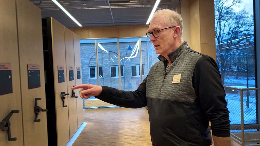 En man pekar mot en av flera skåp i en modern byggnad med stora fönster, som ger en utsikt mot utsidan.
