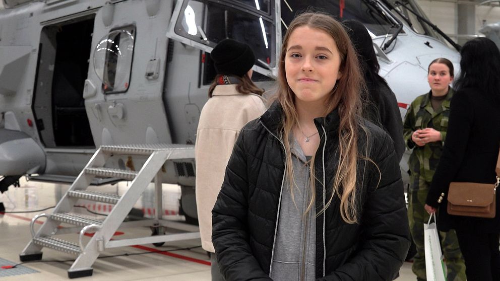 En grupp personer står framför en grå militärhelikopter inomhus. Helikoptern har öppen dörr och en stege som leder upp till den. En person bär en camouflagedräkt.