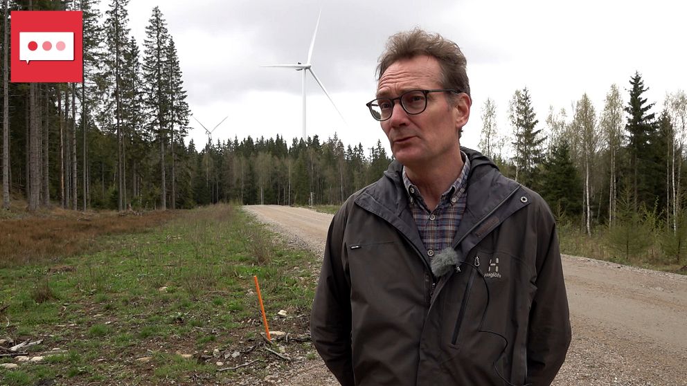 Man på landsväg i skog, med vindkraftverk synligt i bakgrunden.