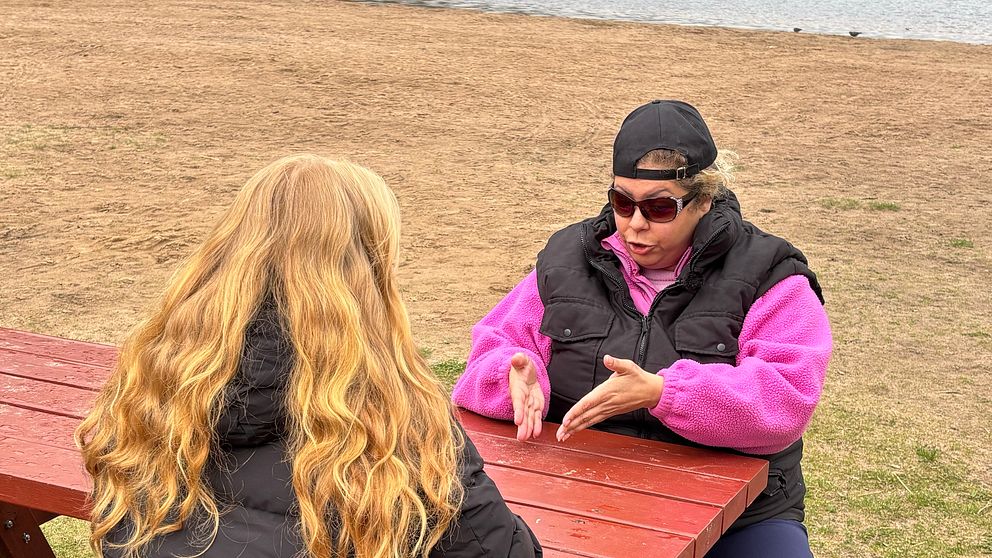 Två personer samtalar vid ett rött picknickbord utomhus nära vatten.
