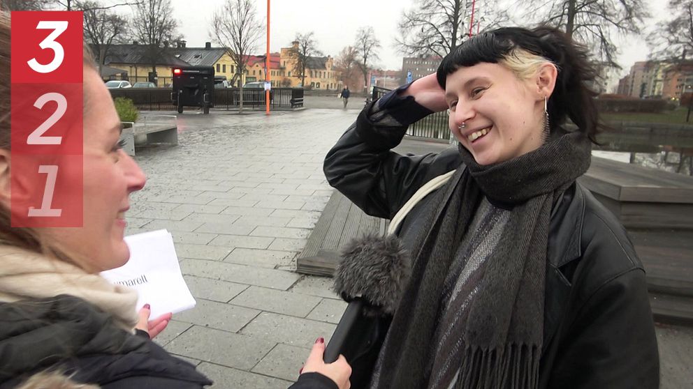 En person med en mörk jacka och en halsduk sträcker sig efter sitt hår medan en annan person, som håller i en mikrofon, ställer frågor. Scenen utspelar sig utomhus på en grå och molnig dag, med träd och byggnader i bakgrunden.