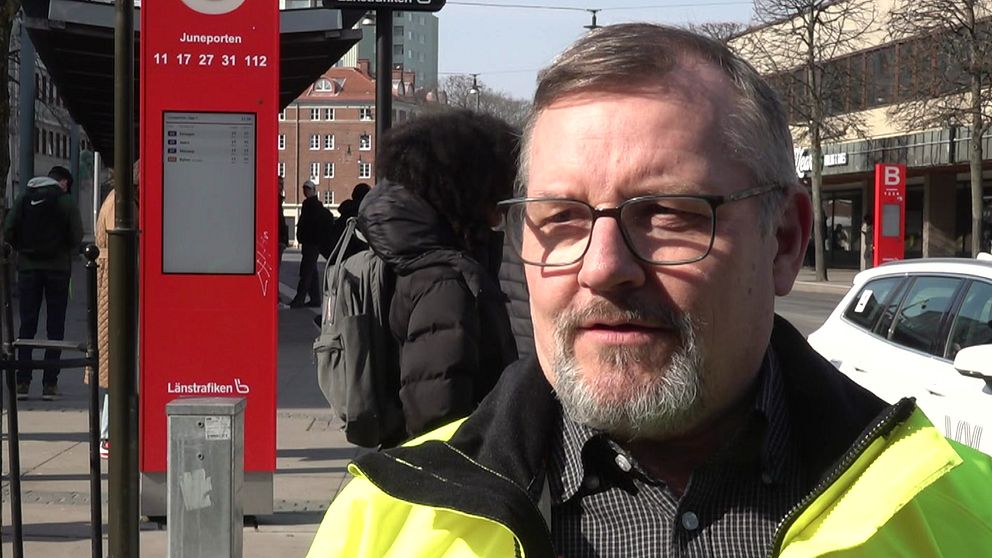 En person i gul reflexjacka vid busshållplats, passerande människor i bakgrunden.