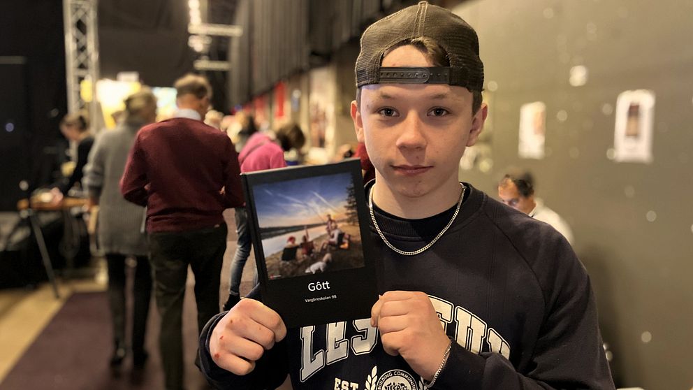 En person håller upp en bok med titeln ”Gött”, som har en bild av en strand på omslaget. I bakgrunden syns en folksamling på ett evenemang.
